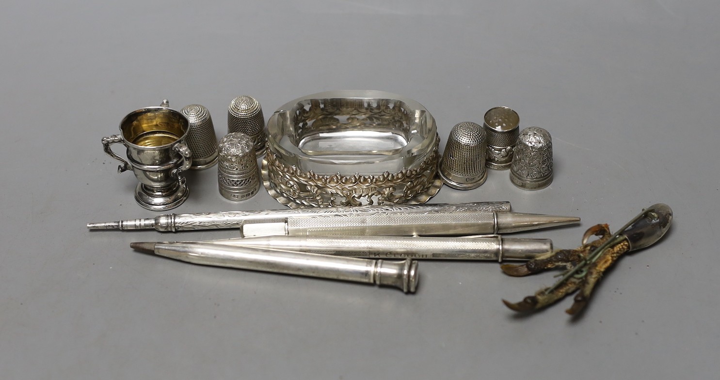 An Edwardian repousse silver mounted glass salt,69mm, a small silver tri-handled trophy cup, five silver thimbles, a plated thimble, three silver cased pens, a sterling pen, one other white metal pen and a brooch.